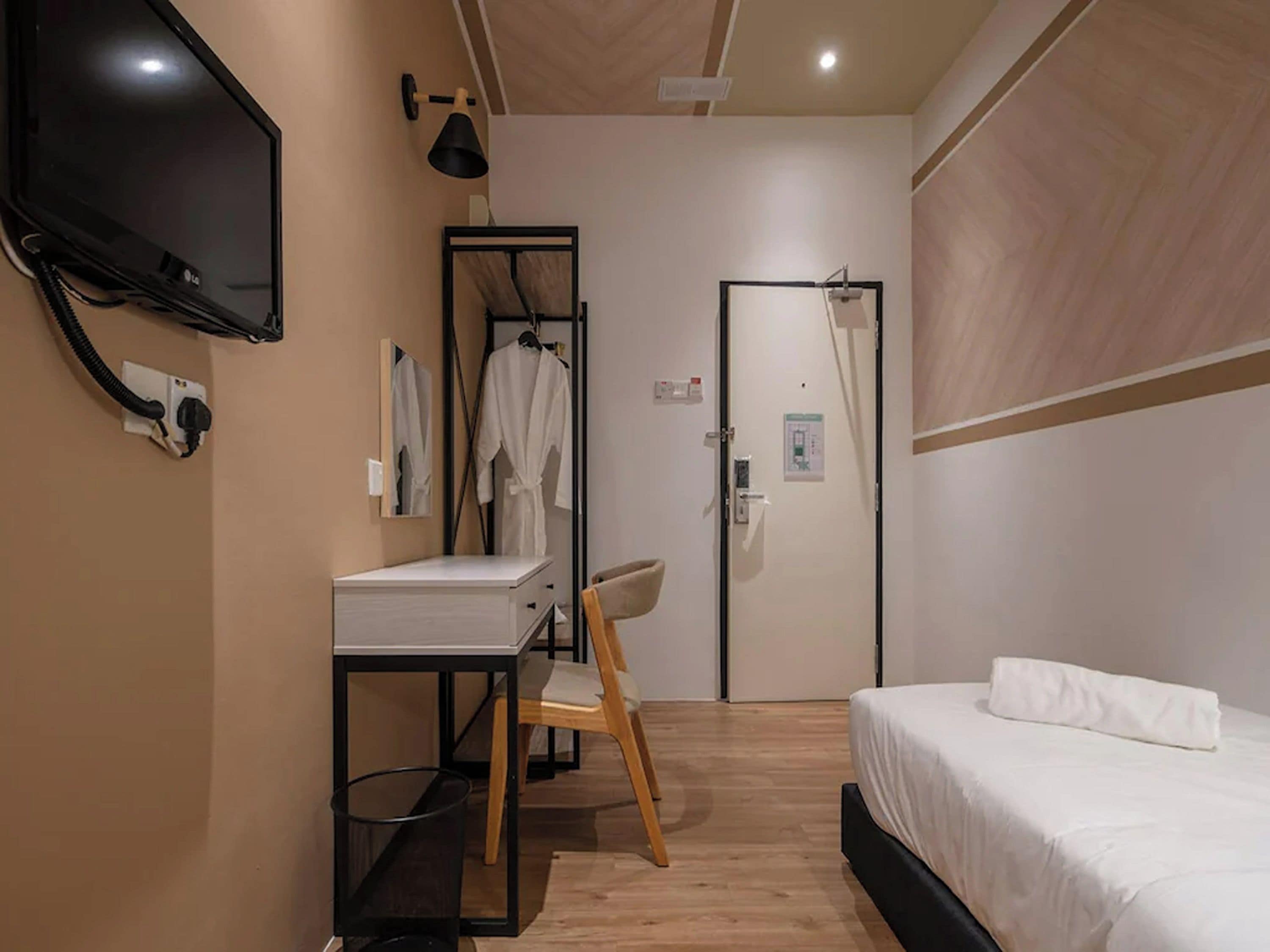 A hotel room with a bed, a desk with a chair, a TV mounted on the wall, and a door with a towel rack.