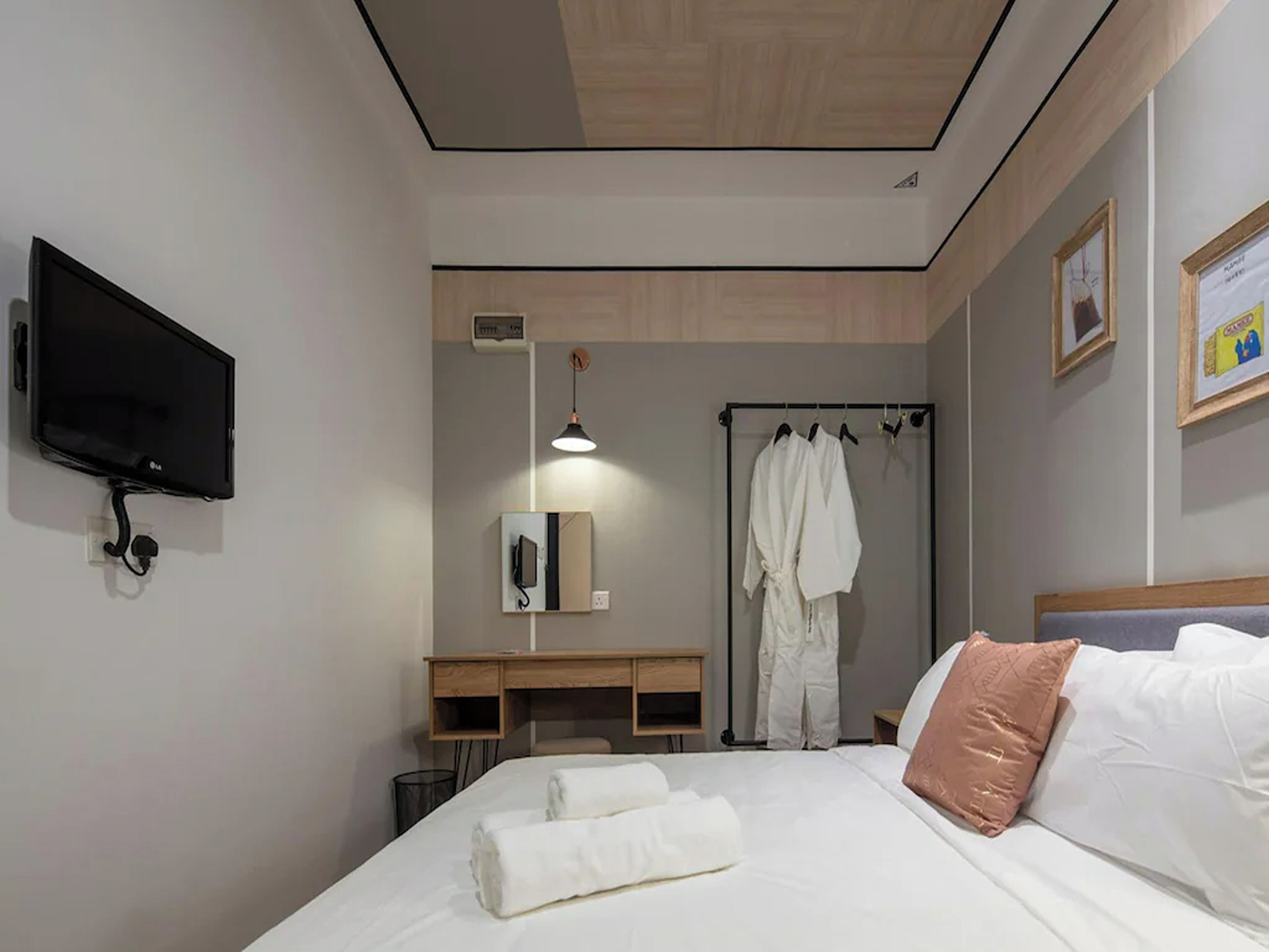 A hotel room with a bed, a TV mounted on the wall, a desk with a chair, a robe rack with robes, and a bedside table.