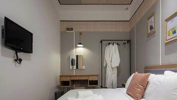 A hotel room with a bed, a TV mounted on the wall, a desk with a chair, a robe rack with robes, and a bedside table.