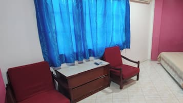 A room with a bed, a desk with drawers, two red chairs, and a blue curtain.