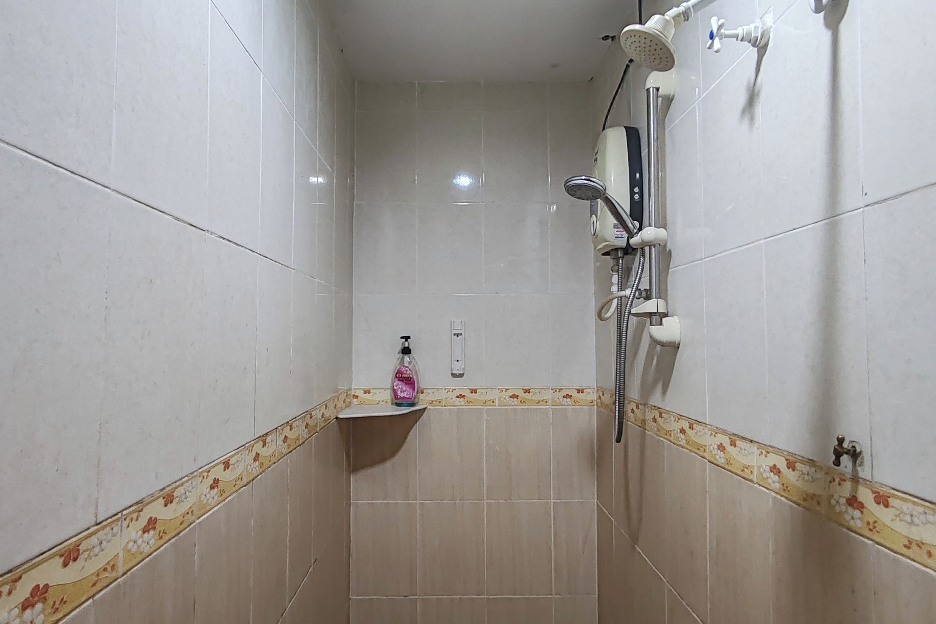 Un piccolo bagno con rivestimento in piastrelle, un soffione manuale, un rubinetto a parete e un dispenser di sapone su uno scaffale.
