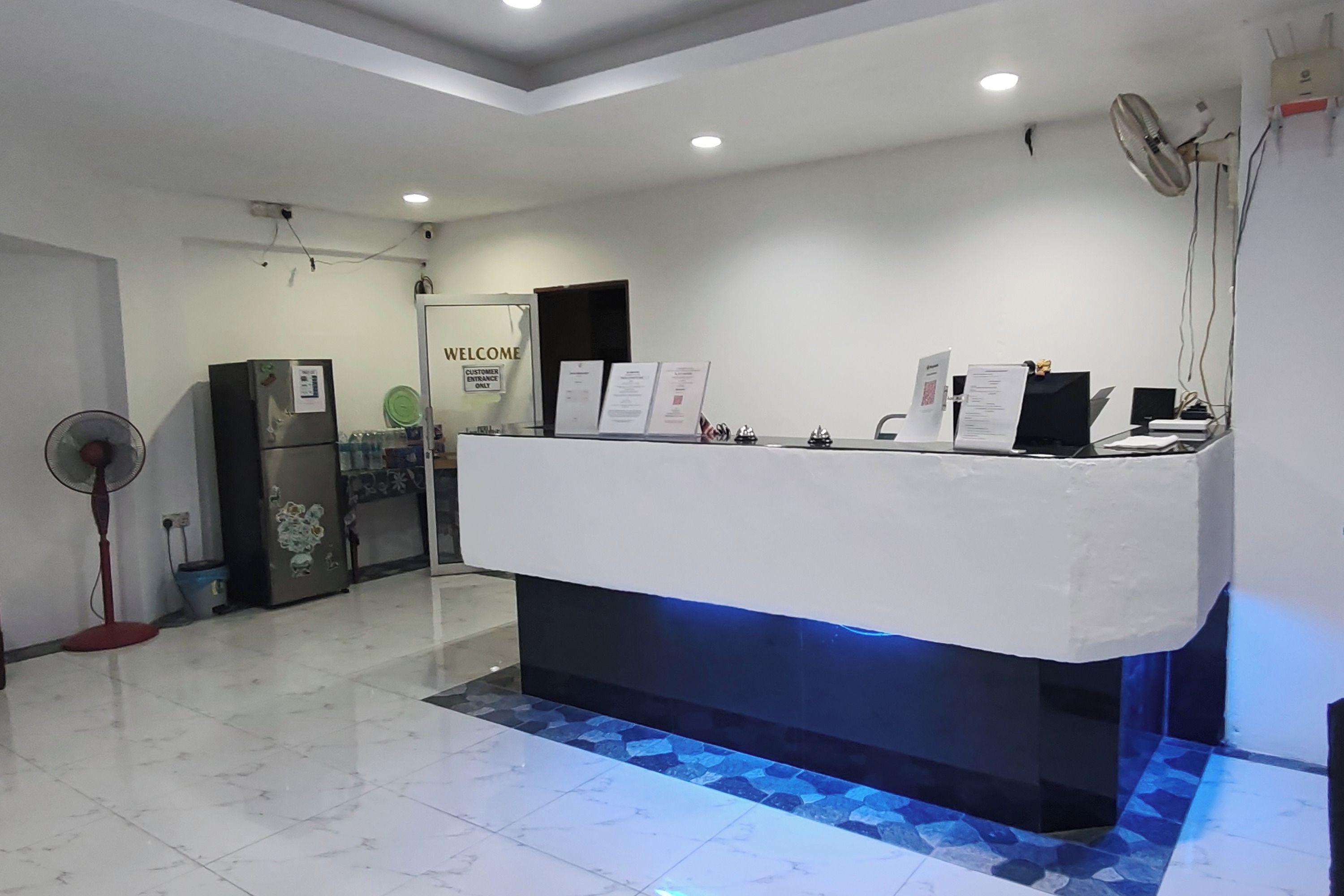 Una reception con base illuminata di blu, un frigorifero, una ventola e un cartello con l'indicazione dell'ingresso clienti.