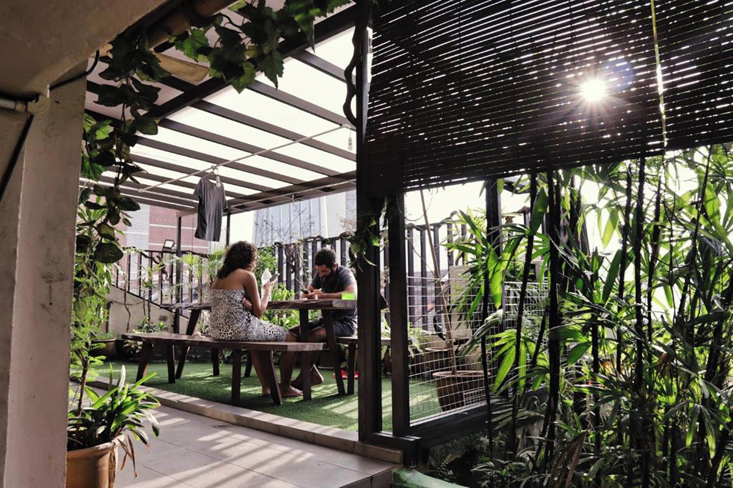 An outdoor seating area with tables and chairs, surrounded by greenery and a pergola structure.