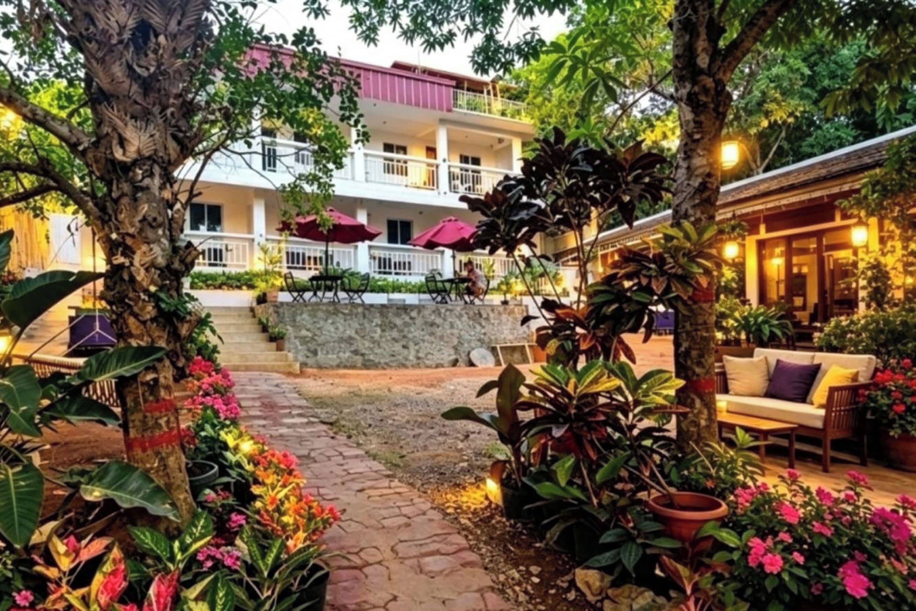 A courtyard with a stone pathway, potted plants, outdoor seating, and a building with balconies and umbrellas.