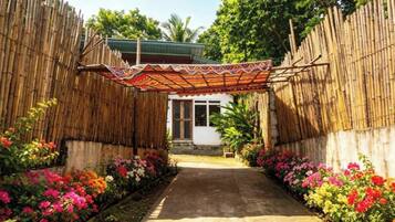 A pathway leading to a building with a thatched roof, surrounded by bamboo walls and colorful flowers.