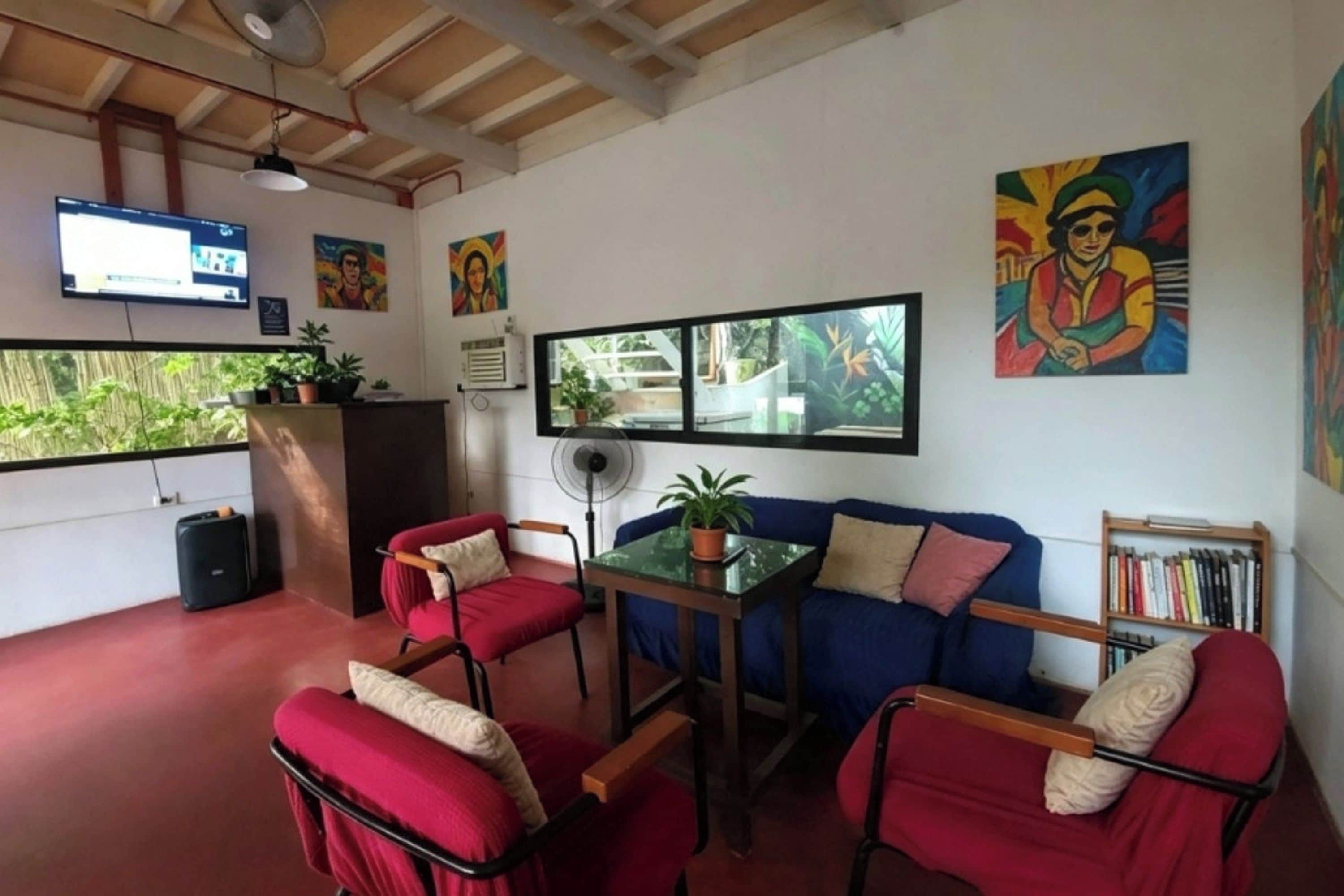 Una habitación con un sofá rojo, dos sillas rojas, una mesita con una planta, un televisor montado en la pared y un ventanal con vistas a la vegetación.