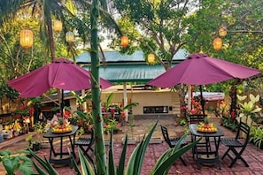 Uma área de estar externa com mesas e cadeiras, rodeada por vegetação exuberante e lanternas decorativas.