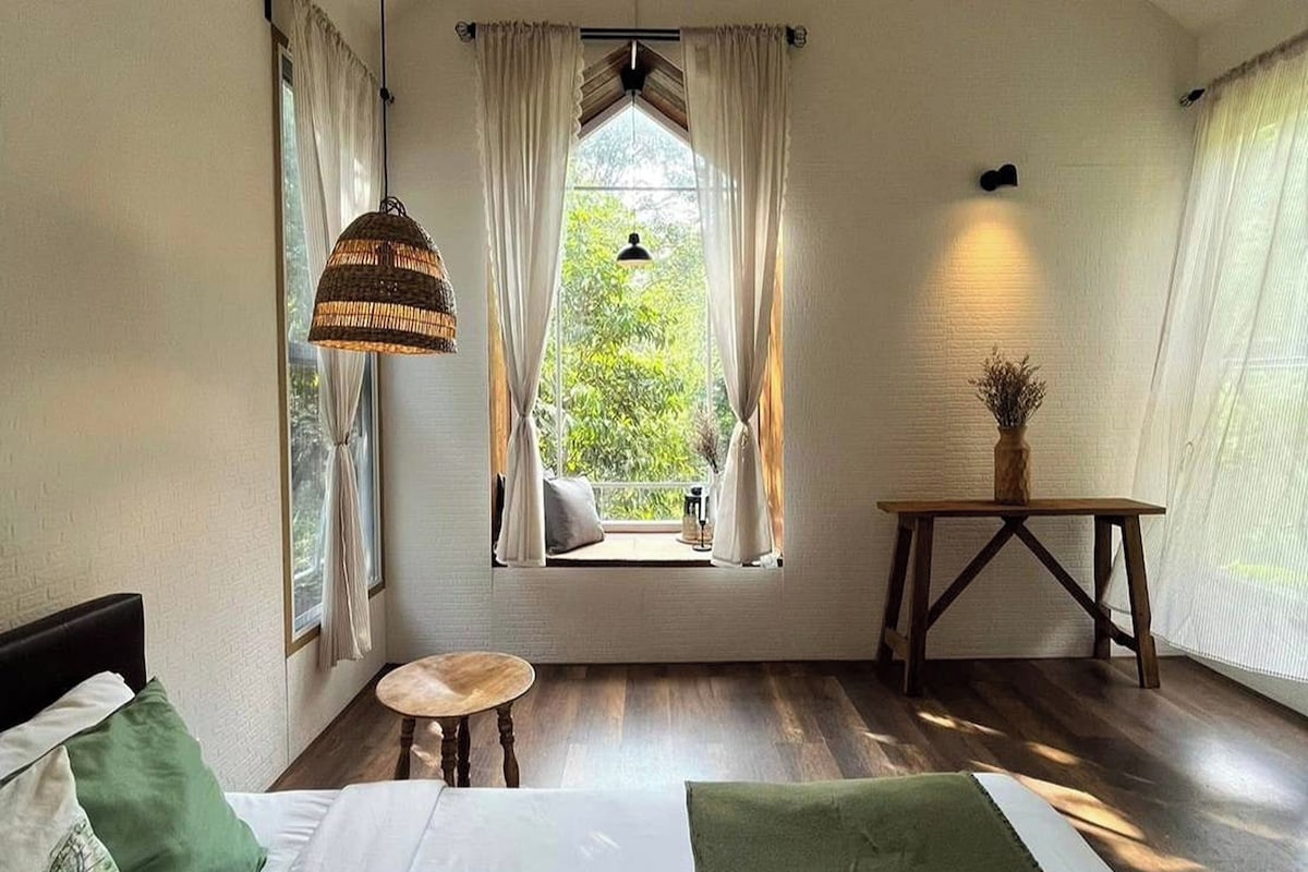 A room with a bed, a small table, a window with curtains, and a hanging light fixture.