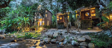 A riverside lodge with multiple wooden cabins, surrounded by lush vegetation and a flowing stream.