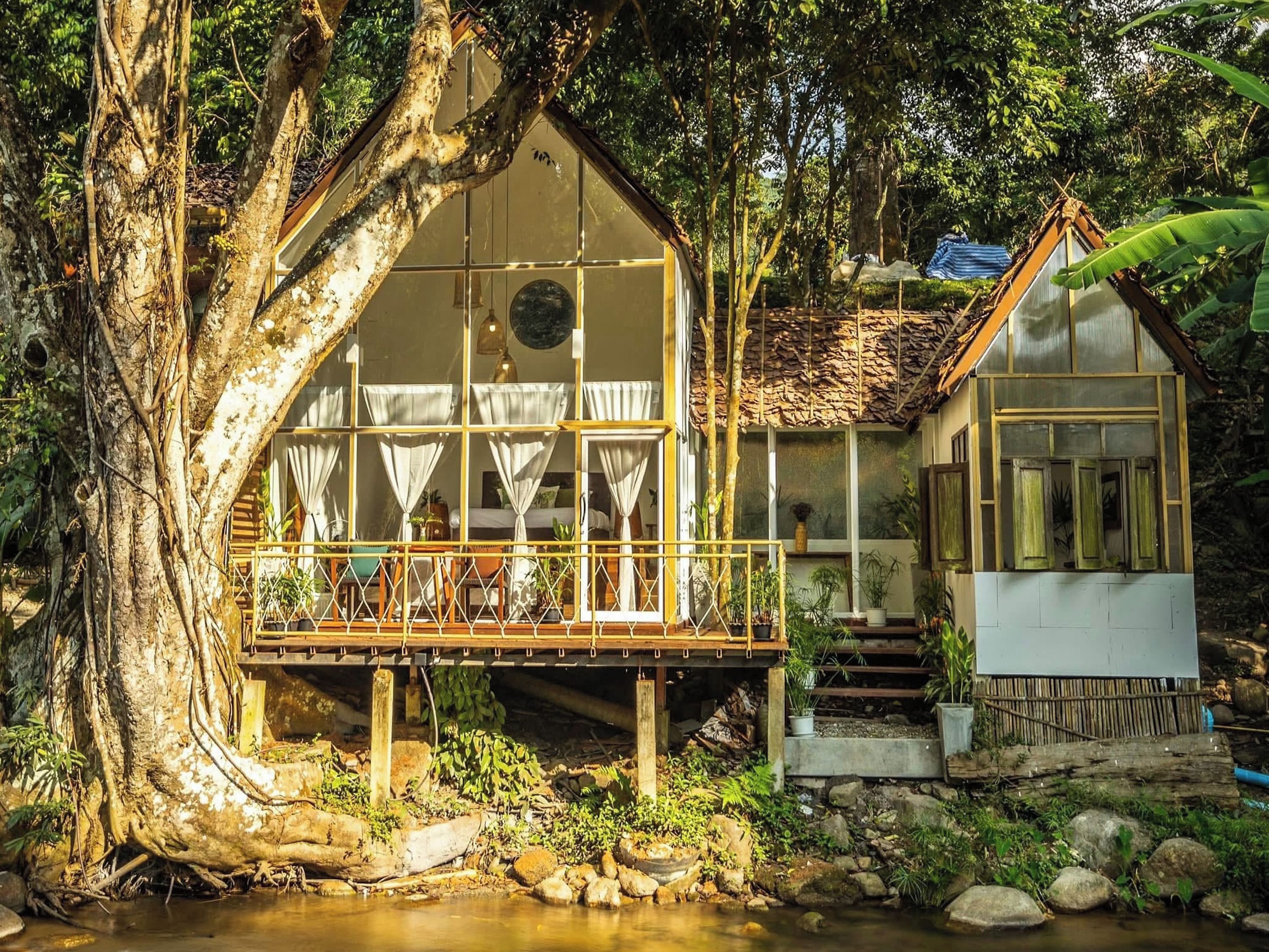 A rustic house with a wooden deck, surrounded by trees and a small stream.