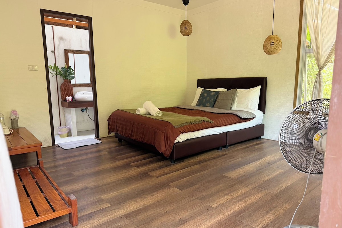 A bedroom with a wooden floor, a bed with a brown headboard, a bedside table, and a fan.