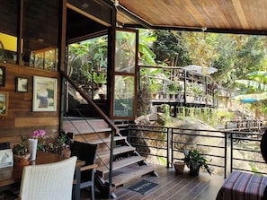 A wooden staircase leading to a balcony with a view of a garden and a small outdoor seating area with a table and chairs. - The Forrest Peace Forest (Mae Kampong) (Chiang Mai)