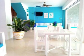 Una habitación luminosa con una mesa y sillas blancas, un ventilador de techo y una pared azul decorada con obras de arte.