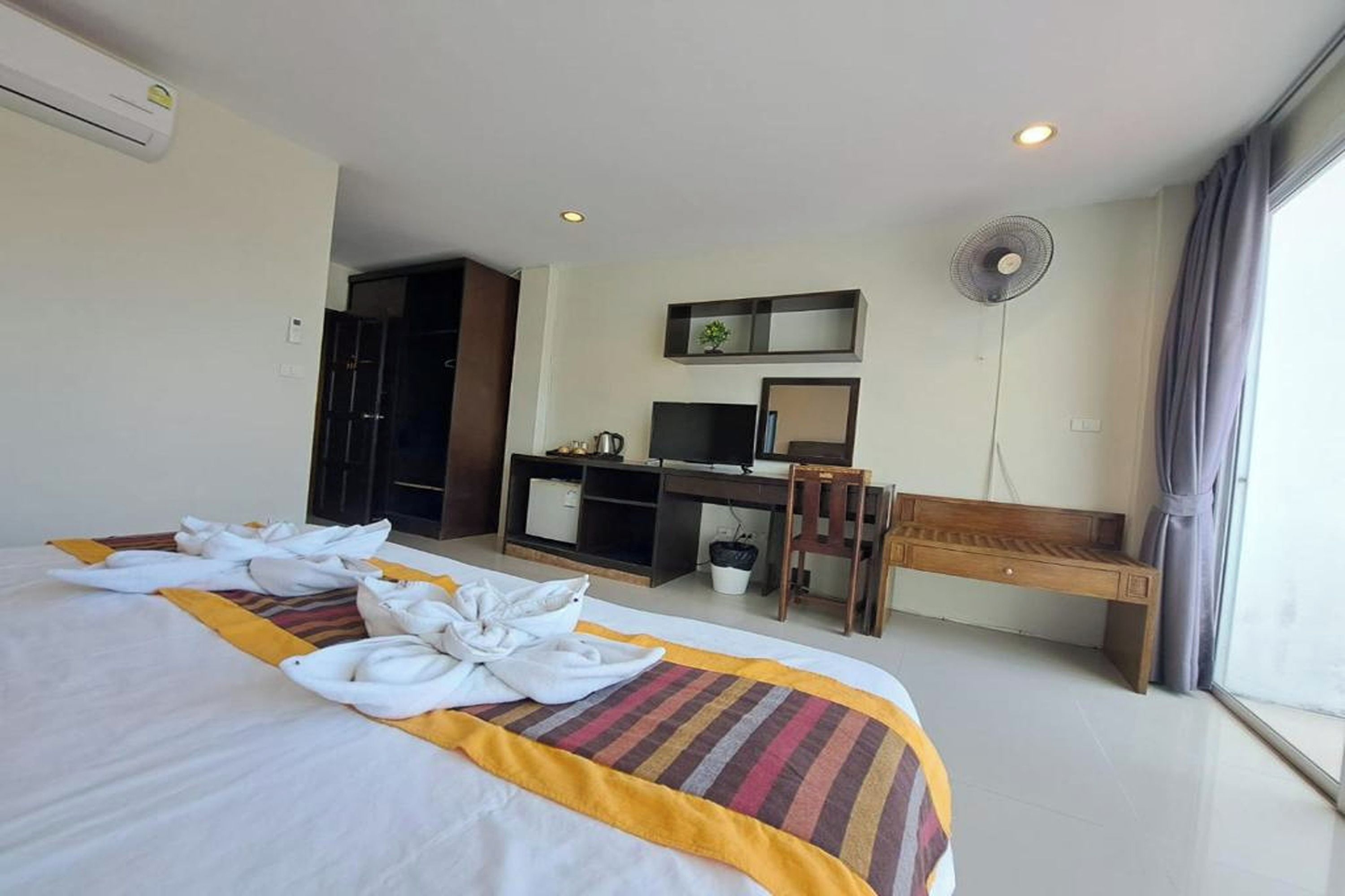Habitación de hotel con cama, escritorio, silla, televisor, ventilador y ventana.