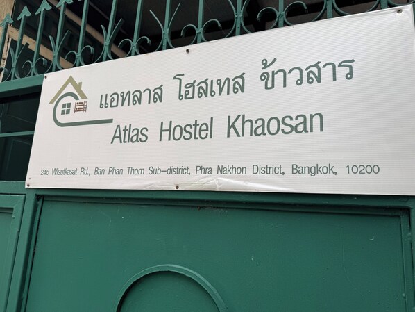 A sign for Atlas Hostel Khaosan with a house icon, indicating it is a hostel. The sign includes the address: 246 Wisutkasat Rd., Bang Phan Thom Sub-district, Phra Nakhon District, Bangkok, 10200. - Atlas Hostel Khaosan (Bangkok)
