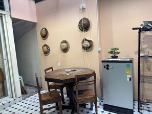 A room with a round wooden table, four chairs, a refrigerator, and several woven hats on the wall. - Atlas Hostel Khaosan (Bangkok)