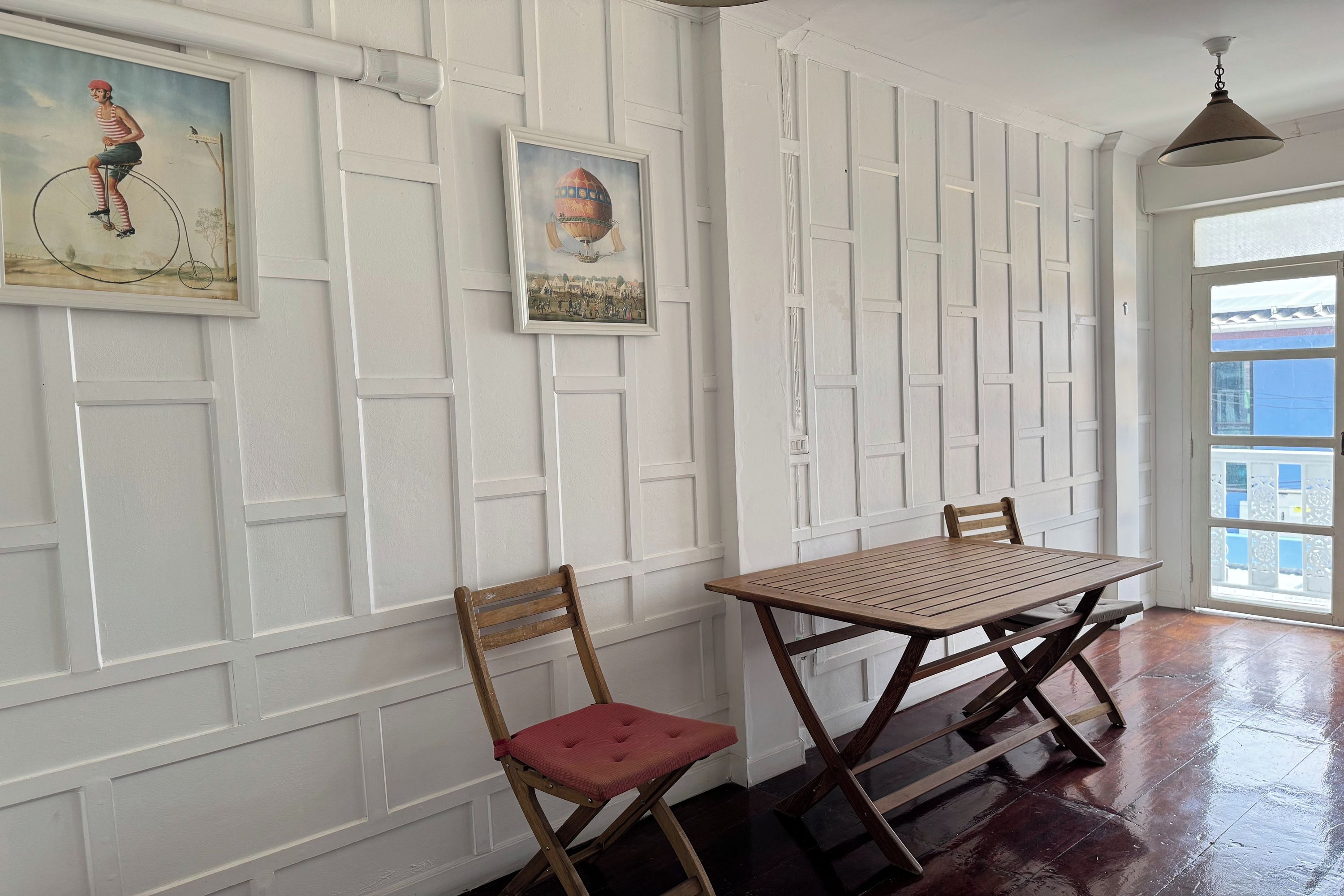 A dining area with a table and chairs, a painting on the wall, and a view through a window.