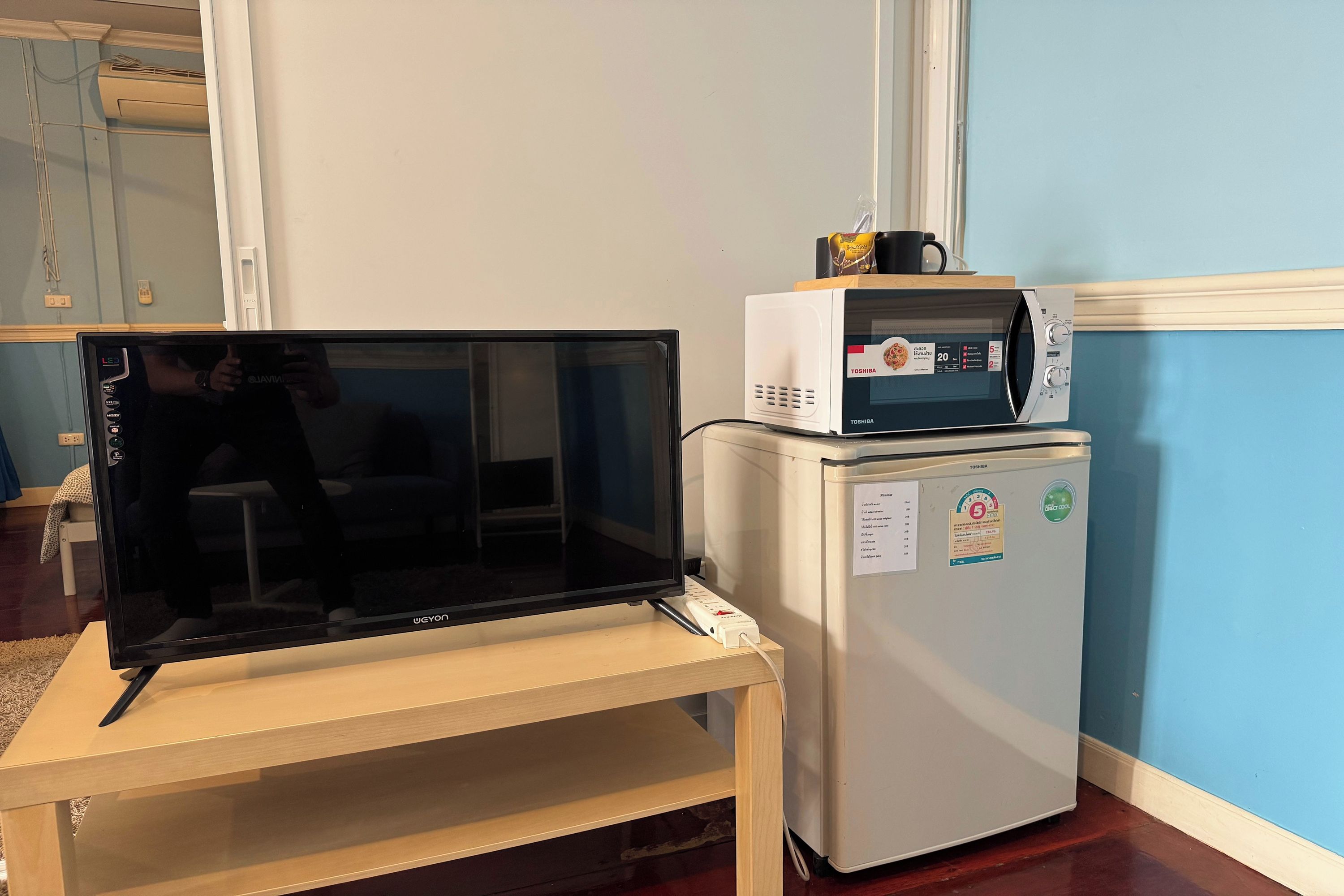 A room with a large flat-screen TV on a stand, a white refrigerator with a microwave on top, and a wall-mounted air conditioning unit.