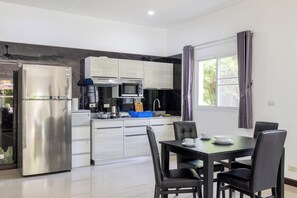 A modern kitchen with a refrigerator, microwave, and stove, a dining table with chairs, and a window with curtains. - Villa Ras Kamala (Kamala)