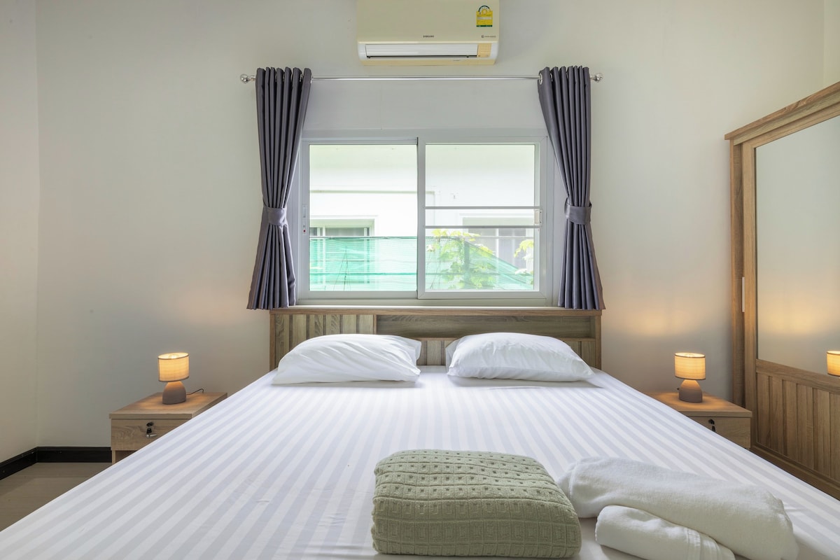 A hotel room with a large bed, two bedside lamps, a window with curtains, an air conditioning unit, and a wooden headboard.