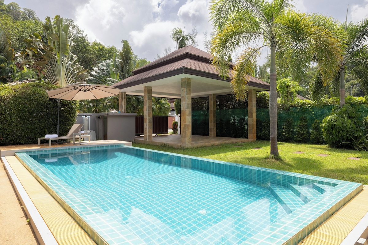 A swimming pool with a tiled deck, a covered patio area, and lush greenery surrounding the property.