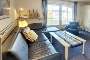 Ein Zimmer mit einer schwarzen Ledersofa, einem Tisch mit einer blau-weißen Tischläufer, einem Schiffmodell auf einem Regal und einem Stuhl mit blauer Sitzkissen.
