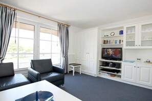 A living room with a sofa, a small table, a television, and white cabinets with glass doors. - Holiday apartment with 1 bedroom (Wyk auf Föhr)