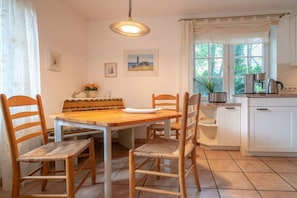 A dining area with a wooden table and chairs, a hanging light fixture, a window with curtains, and a kitchen area with cabinets and appliances. - Holiday apartment with 1 bedroom (St.Peter-Ording)