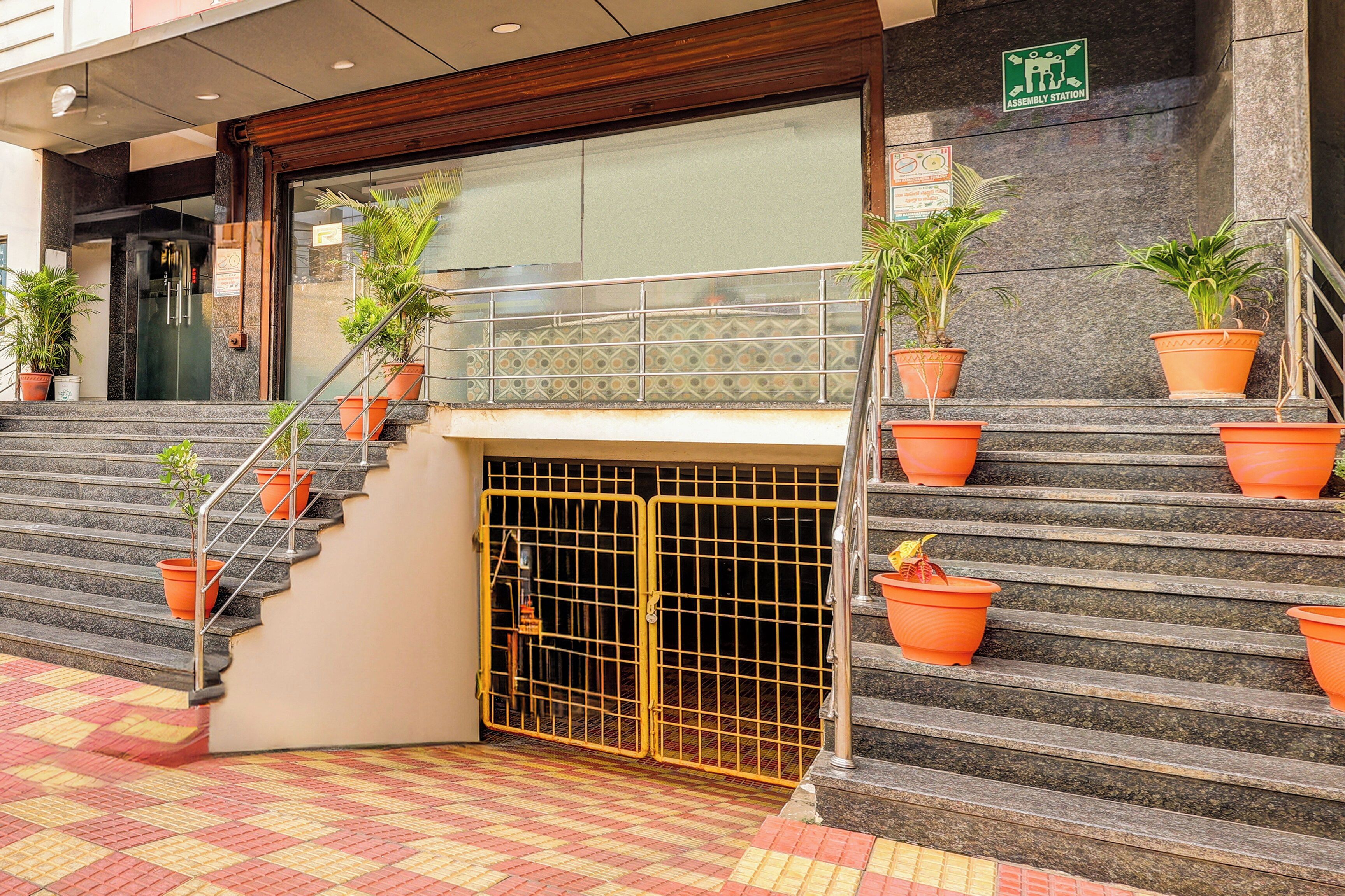 Entrada de hotel con escaleras, plantas en macetas y señalización de punto de encuentro de emergencia.