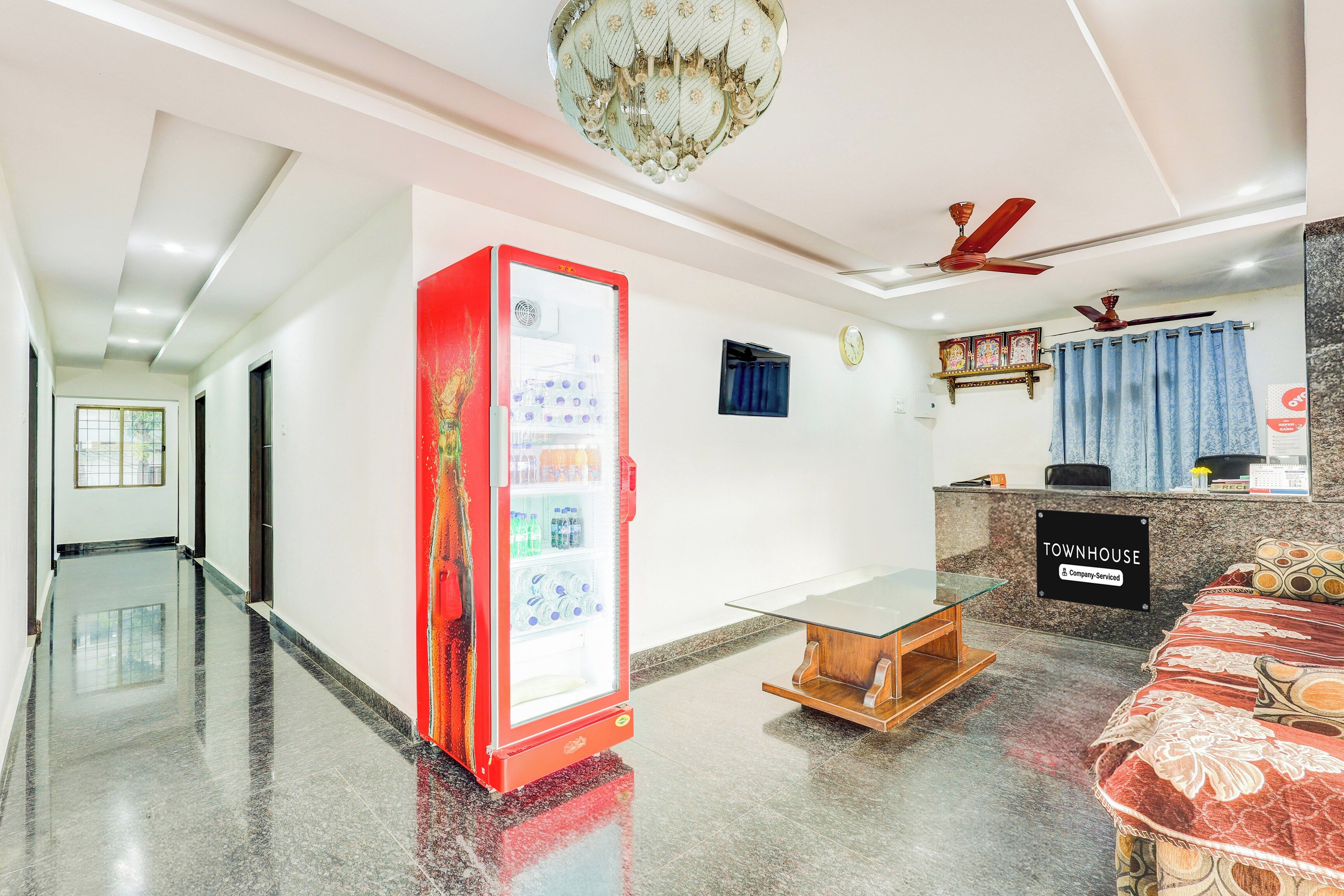 Un vestíbulo de hotel con mostrador de recepción, un refrigerador lleno de bebidas, un reloj y un ventilador de techo.