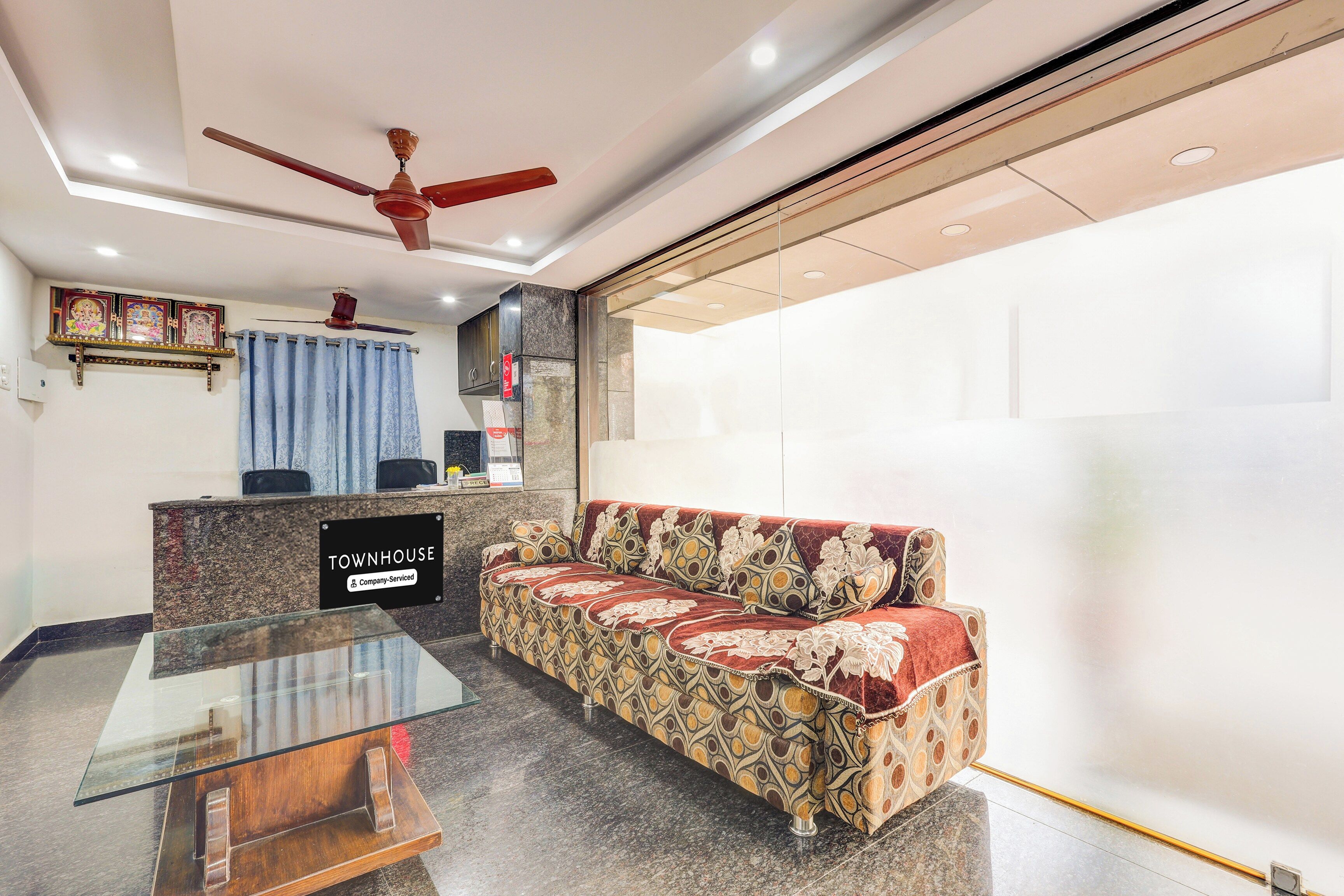 Un área de recepción con un sofá, una mesa de vidrio, un ventilador de techo y un letrero que indica que el establecimiento es de servicio corporativo.