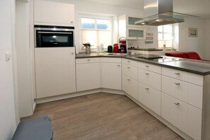 A modern kitchen with white cabinetry, a built-in oven, a stainless steel range hood, and a red chair. - Strandstraße 18 b, Apt. 1, Haus Paula (Wyk)