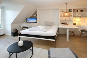 A modern bedroom with a bed, a desk, and a chair. There is a television on a shelf, a lamp, and a bookshelf. - Lüttmarsch 13, Holiday Apartment 10 (Wyk auf Föhr)