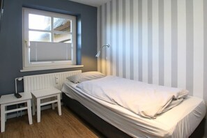 Ein Schlafzimmer mit einem Einzelbett, einem Nachttisch, einem Sessel, einem Fenster mit Jalousien und einer Heizung.