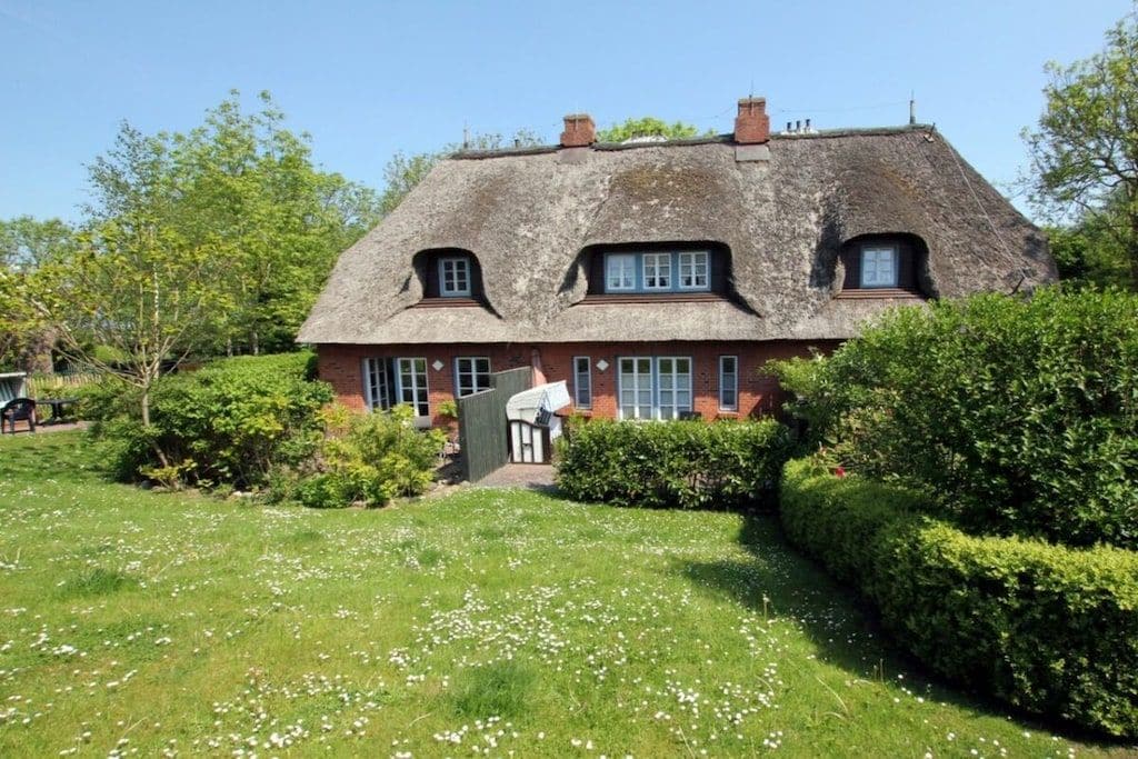 Une maison traditionnelle à toit de chaume, dotée d’une façade en briques, entourée d’un jardin bien entretenu avec une pelouse et des haies.