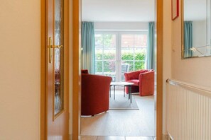Ein Blick ins Zimmerinnere zeigt eine Holztür, einen roten Sessel, einen weißen Tisch und durch eine Glastür einen Ausblick auf eine grüne Außenanlage.