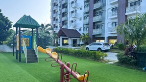 Ein Spielplatz mit Wippe und Klettergeräten, umgeben von einem gepflegten Rasen und einem Wohnhaus mit Balkonen.