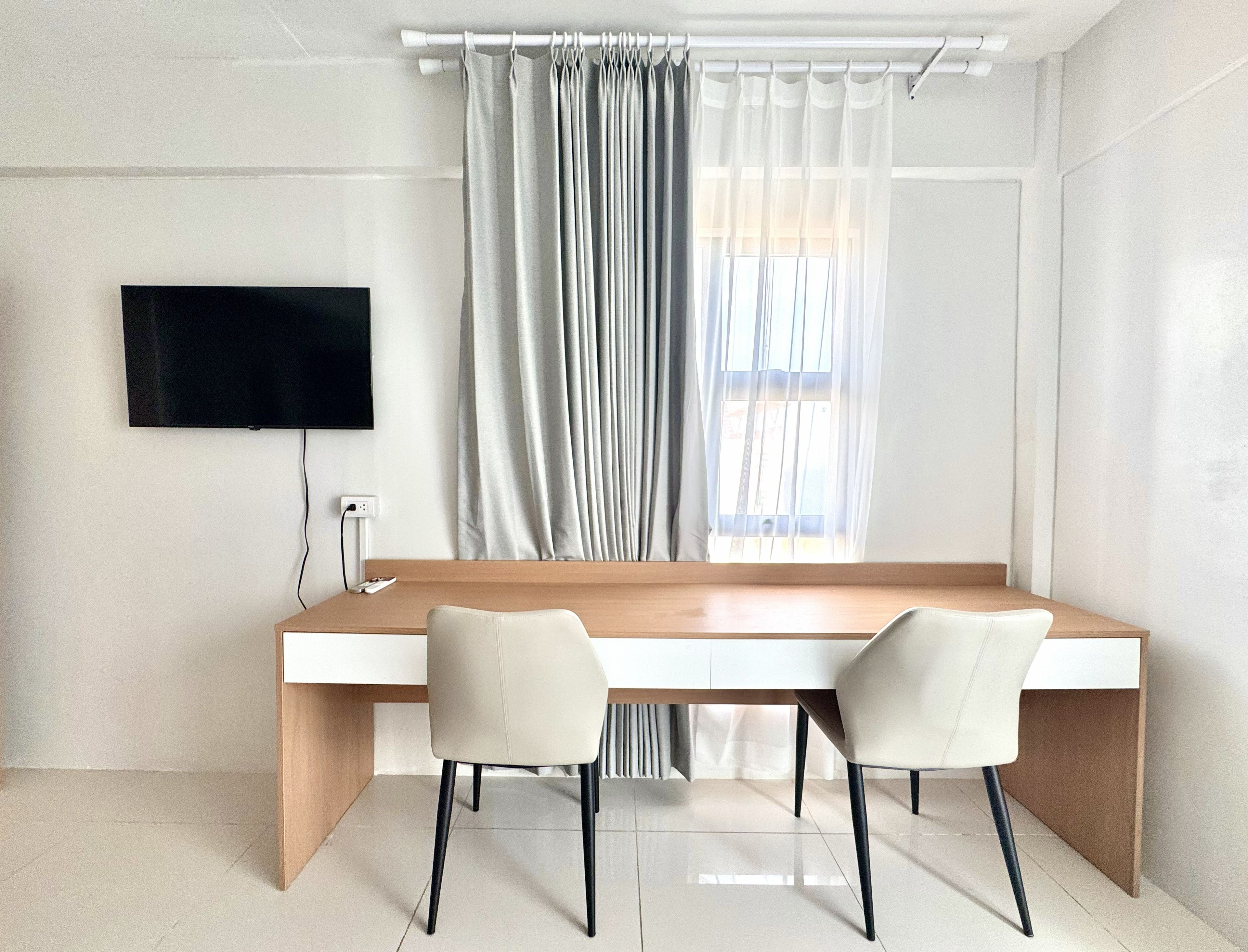 Ein modernes Zimmer mit einem langen Holztisch, zwei Stühlen, einem an der Wand befestigten Flachbildfernseher und einem Fenster mit durchsichtigen Vorhängen.