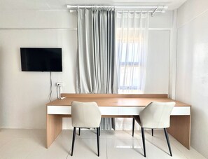 Una habitación moderna con un escritorio de madera, dos sillas, un televisor de pantalla plana en la pared y un ventanal con cortinas traslúcidas.
