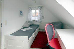 Ein kleines Zimmer mit einem Einzelbett, einem Schreibtisch, einem Stuhl, einem Heizkörper und einem fenster mit Vorhängen.