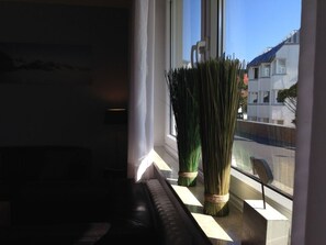 A window with a view of buildings, a black leather sofa, and a tall plant with a brown base. - Holiday Apartment With 1 Bedroom (Norderney)