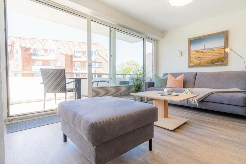 A modern living room with a grey sofa, a wooden coffee table, and a large window offering a view of residential buildings.