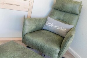 A green upholstered armchair with a cushion labeled 'Lieblingsplatz' in a room with a wooden floor and a light-colored wall. - Holiday apartment with 2 bedrooms (St. Peter-Ording)