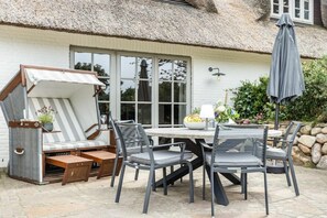 An outdoor patio with a thatched roof, a dining table with chairs, and a seating area with a cushioned bench. - Holiday home Captain Karl Keitum / Sylt (Nordfriesland)