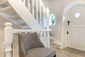 A staircase with white balusters, a grey upholstered chair, and a white door with a window above it. - Holiday home with 1 bedroom (Nordfriesland)
