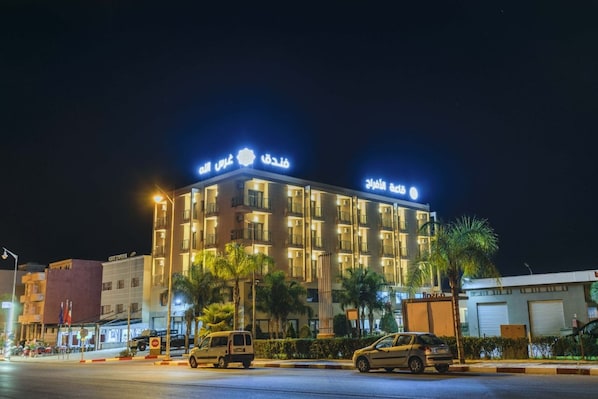 Un hotel de varios pisos con señalización iluminada, un estacionamiento y vista a la calle de noche.