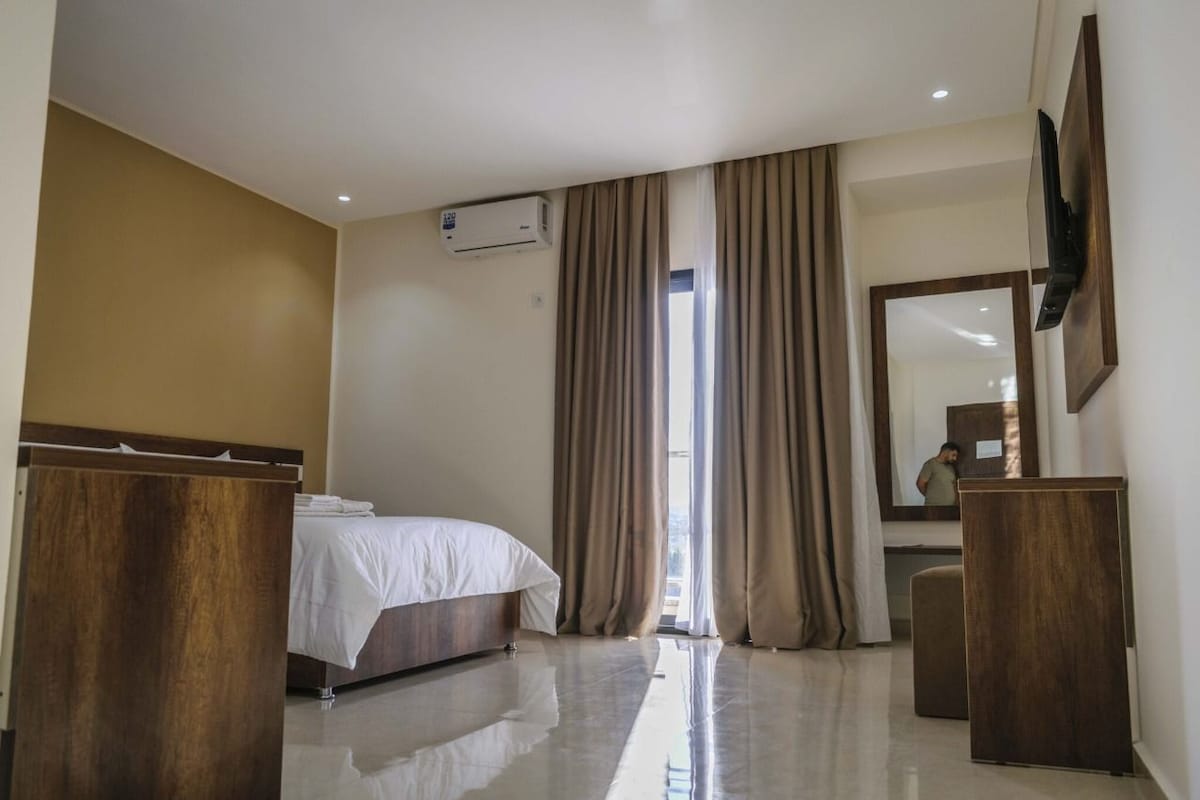 A hotel room with a bed, a wooden dresser, a television mounted on the wall, and an air conditioning unit.