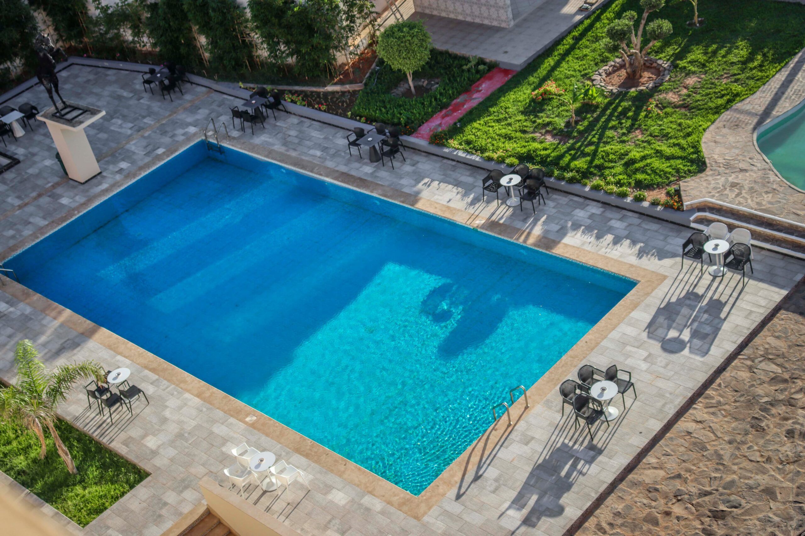 A rectangular swimming pool with clear blue water, surrounded by stone tiles and outdoor seating areas with tables and chairs.