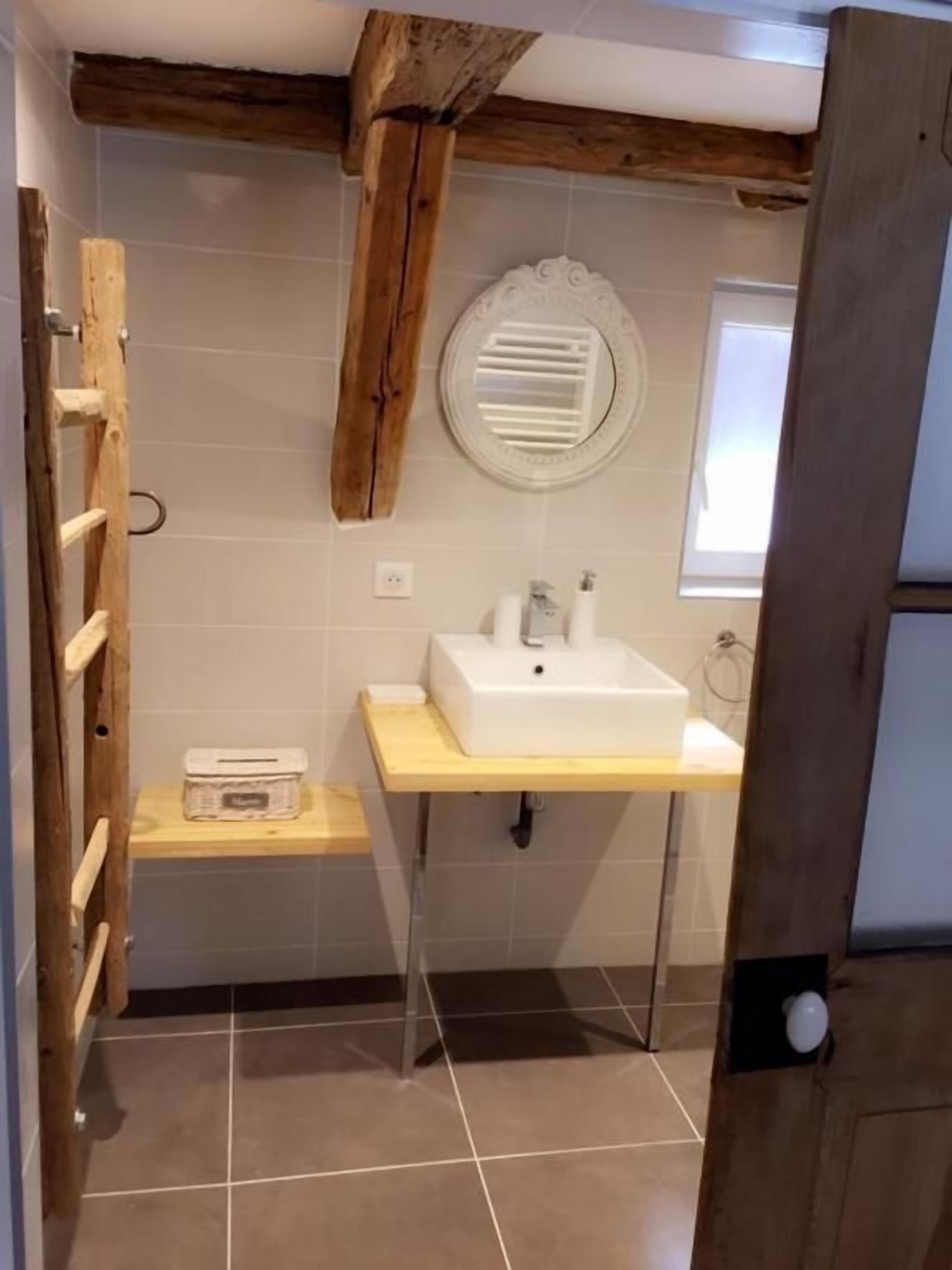 Een badkamer met een plafond van houten balken, een wit wastafelmeubel, een spiegel en een raam.