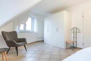Ein Zimmer mit großem Schrank, einem Sessel, einem kleinen Tisch und einem Fenster mit Vorhängen.